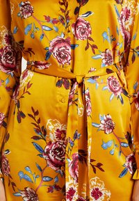Gul blommig klänning med röda och blå mönster, tillverkad av mjukt tyg. Har knäppning med knappar och ett knytbart midjeband för en figurnära passform.