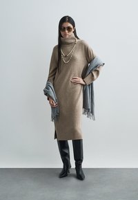 Barna kötött garbóruha hosszú ujjakkal, oldalsó slicccel. Szürke rojtos sállal és fekete térdig érő csizmával párosítva, gyönggyel díszített nyaklánccal kiegészítve.