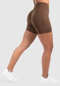 Pantaloni corti marroni a vita alta con una vestibilità aderente, texture liscia e tasche laterali. Indossati con scarpe da ginnastica bianche.