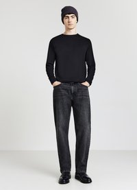 Maglietta nera a maniche lunghe, vestibilità classica; jeans di denim grigio scuro con una leggera scoloritura; berretto scuro; scarpe nere; sfondo semplice.