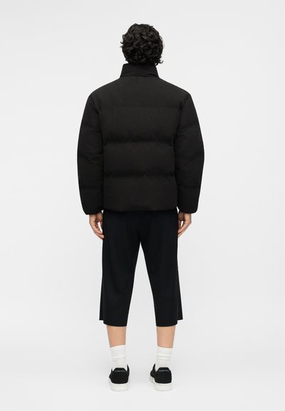 Άνδρας με σγουρά μαλλιά φορώντας μαύρο μπουφάν με επένδυση, μαύρο παντελόνι μέχρι τον αστράγαλο, λευκές κάλτσες και μαύρα αθλητικά παπούτσια, στέκεται γυρισμένος προς τα πίσω.