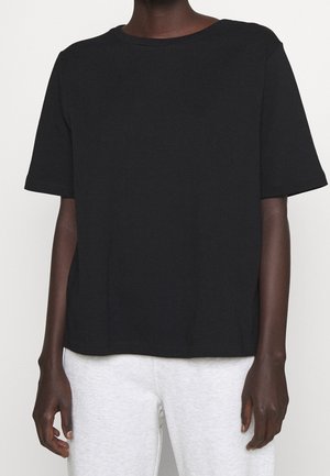 Person iført en ensfarvet sort kortærmet t-shirt og lysegrå bukser, stående mod en lys baggrund.