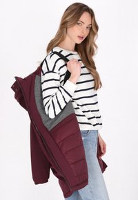 Burgundska jakna s preklopom i sivim naglascima, prikazana otkopčana. Model nosi prugastu košulju dugih rukava, plave traperice i torbu.