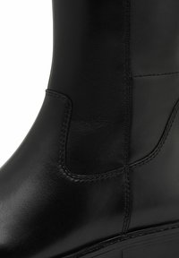 Botte en cuir noir avec une texture lisse, des coutures latérales et un design structuré. Tige haute et élégante avec une légère courbe.