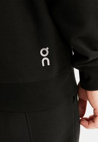 Black sweatshirt with ribbed cuffs and hem. Features a silver logo on the lower side, highlighting its simple and functional design.
