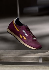Baskets Reebok bordeaux avec des rayures jaunes et une semelle noire flottant au-dessus d'escaliers en béton dans un éclairage tamisé.