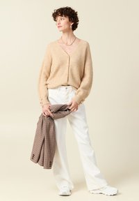 Gilet tricoté beige avec un décolleté en V, des boutons sur le devant et des manches texturées. Associé à un pantalon large blanc et tenant une écharpe à carreaux.