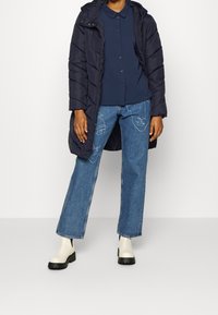 Mörkblå puffjacka med huva över en mörkblå skjorta med krage, i kombination med ljusblå jeans med abstrakta ansiktslinjer och vita ankelskor.