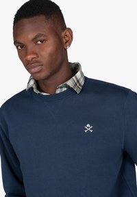 Joven con un pendiente de diamante, sudadera azul marino con un pequeño logo blanco y el cuello de una camisa de cuadros visible por encima del escote.