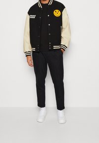 Černá a krémová bomber bunda s žebrovanými rukávy a límcem, s žlutou nášivkou s úsměvem a logem "Tommy Jeans" na hrudi.