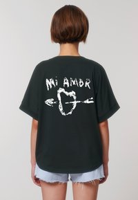 Een persoon met kort haar draagt een donker t-shirt met de tekst "Mi Amor" en een hart met een pijl, in combinatie met denim shorts.