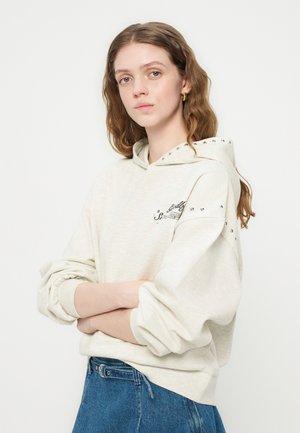 Młoda kobieta z długimi falowanymi włosami, ubrana w kremową bluzę z kapturem z metalowymi ćwiekami i niebieskie dżinsy, stoi z założonymi ramionami na białym tle.