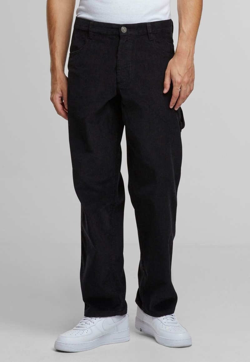 Homme portant un pantalon en velours côtelé noir, des baskets blanches et une chemise blanche, debout devant un fond gris uni.