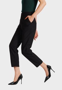 Pantaloni neri su misura con vestibilità slim e lunghezza alla caviglia, dotati di tasche laterali e un tessuto liscio. Indossati con tacchi a spillo in pelle lucida nera.