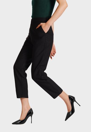 Pantaloni neri su misura con vestibilità slim e lunghezza alla caviglia, dotati di tasche laterali e un tessuto liscio. Indossati con tacchi a spillo in pelle lucida nera.