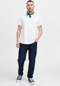 Valkoinen lyhythihainen t-paita, tummansiniset farkut, mustat kankaiset tennarit. Teal-värinen bandana kaulassa, seisoo neutraalin taustan edessä.