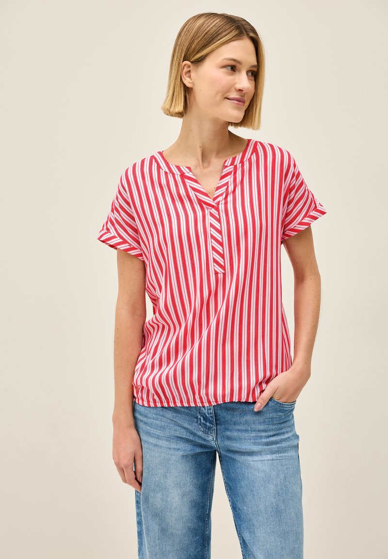 Rood-wit gestreepte blouse met korte mouwen, een v-hals, een ontspannen pasvorm en een elastische onderrand. Gecombineerd met lichtblauwe denim jeans.