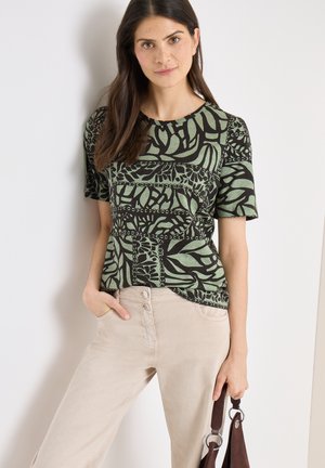 Femme portant un haut à manches courtes vert et noir à motifs, et un pantalon beige, tenant un sac à main marron avec des accents métalliques circulaires.