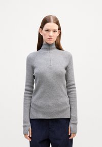 Grauer gerippter Rollkragenpullover mit einem Viertelreißverschluss, langen Ärmeln und tailliertem Design. Getragen mit dunkelblauen Cordhosen.