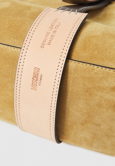 Barna bőrszíjjal ellátott, világosbarna velúrból készült táska, amelyen a "Genuine Leather Made in Italy" és a "Moschino Milano" feliratok barna betűkkel vannak gravírozva.