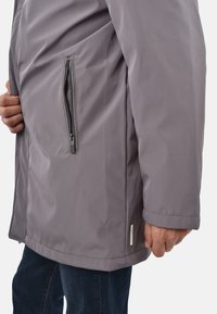 Uomo con giacca grigia con tasca laterale con cerniera, che mostra la mano vicino alla tasca e indossa jeans blu su sfondo uniforme.