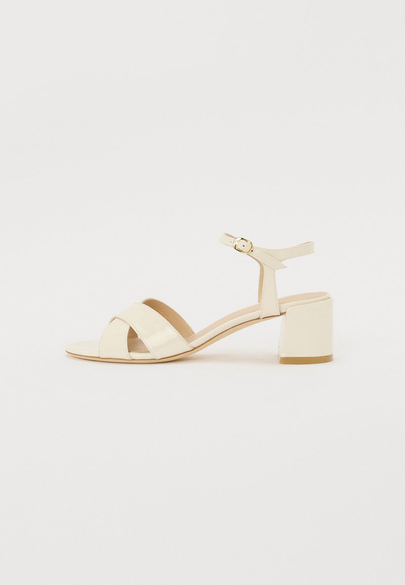 Beige patent leather heeled sandals with cross strap design, open toe, adjustable ankle strap, and a block heel. Smooth texture.