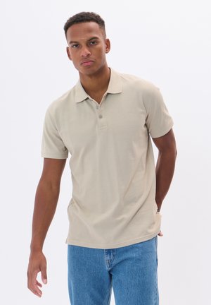 Polo beige en tissu léger, avec un col classique, une patte de boutonnage à trois boutons et des manches courtes. Associé à un jean en denim bleu.