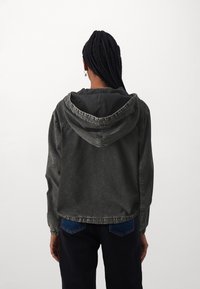 Mörkgrå denimjacka med huva, utrustad med elastiska ärmslut, en avslappnad passform och kontrasterande sömmar. Bakifrån visas huvan form.