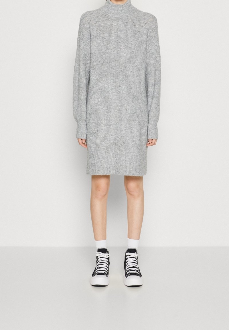 Personne portant une robe-pull en maille grise à col roulé avec manches longues et des baskets montantes noires avec lacets blancs et chaussettes.