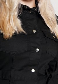Veste noire avec un design à boutonnière, présentant des accessoires en argent, deux poches poitrine et une taille nouée pour un look ajusté. Texture de tissu douce.