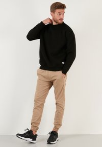 Hanorac negru cu guler rotund și mansete ribbate, asortat cu pantaloni jogger bej cu mansete elastice și buzunare laterale, purtat cu adidași negri.