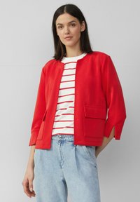 Rode, lichte jas met een ronde hals, driekwartmouwen en twee voorzakken, gedragen over een wit gestreept T-shirt en blauwe jeans.