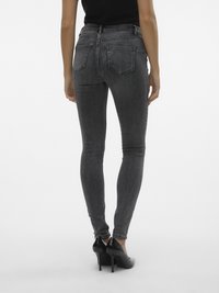 Mujer vestida con jeans ajustados de color gris oscuro y zapatos de tacón negro, de pie con la espalda hacia la cámara contra un fondo blanco.