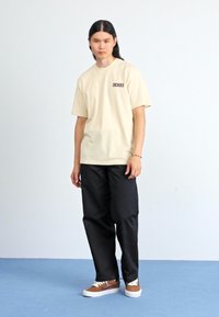 Beige t-shirt med kort ærme og trykt logo, parret med løstsiddende sorte bukser og brune sneakers. Enkel design og afslappet pasform.