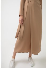 Personne portant un long manteau beige et une jupe, debout avec une main détendue, chaussée de baskets blanches, sur un fond clair uni.