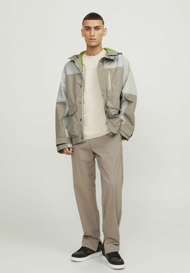 Lichte, grijze en beige jas met een groen accent op de capuchon, gecombineerd met beige broek in een relaxed fit en zwarte sneakers met witte zolen.