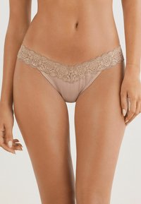String beige avec une délicate ceinture en dentelle, ornée de motifs floraux. Le tissu a une texture lisse et offre un design à couverture minimale.