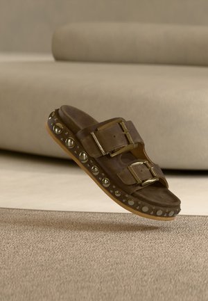 Sandale à glissière en daim marron avec deux grandes boucles dorées et des clous ronds dorés le long du bord de la semelle sur un tapis beige.