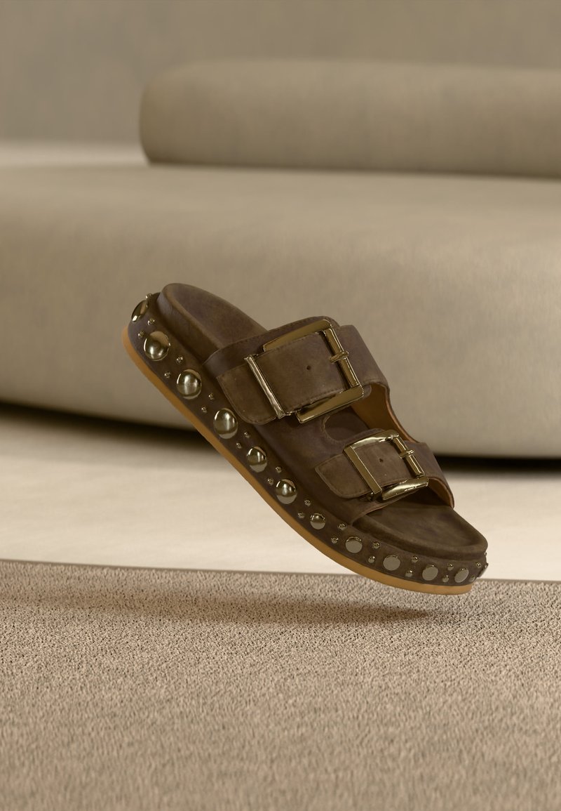 Καφέ σουέντ σανδάλι slide με δύο μεγάλες χρυσές αγκράφες και στρογγυλές χρυσές τρούκς κατά μήκος της άκρης της σόλας πάνω σε μπεζ χαλί.