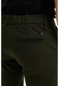 Green patterned trousers with a textured design, featuring a back pocket and a black button accent, made from a woven material.