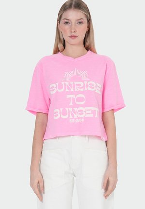 Femme portant un t-shirt court rose clair avec le texte "Sunrise to Sunset Est-2005" et un pantalon blanc, debout sur un fond blanc.
