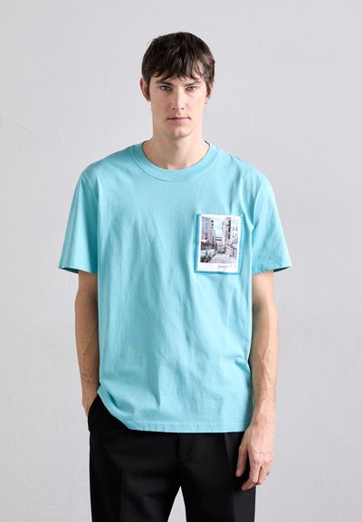 Helles blaues Baumwoll-T-Shirt mit einem rechteckigen Grafik-Patch auf der Brust, das eine Straßenansicht in gedämpften Farben zeigt. Kurze Ärmel, Rundhalsausschnitt.