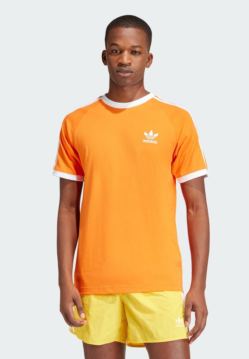 Camiseta de algodón naranja con detalles blancos, cuello redondo y logo de Adidas en el pecho izquierdo. Modelo vistiendo pantalones cortos amarillos.
