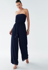 Námorní jumpsuit bez ramínek s plisovanou podprsenkou, elastickým pásem a širokými kalhotami. Doplňuje se zlatými šperky a sandály na vysokém podpatku.