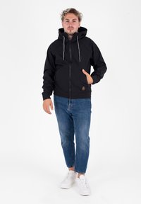 Giovane uomo in piedi con una mano in tasca, che indossa una giacca nera con cappuccio, jeans blu e sneakers bianche su sfondo bianco.