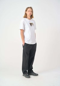 White cotton t-shirt with a playful graphic design, paired with loose black trousers and dark sneakers, standing against a plain background.