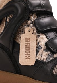 Gros plan d’une basket en cuir noir avec une texture effet peau de serpent et une étiquette en daim beige portant la mention "BRONX", attachée par une chaîne à billes argentée.