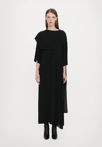 Black long dress with draped front and sleeves, made of lightweight fabric. It features a relaxed fit and is paired with black heeled boots.