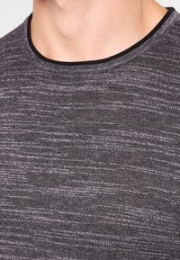 Chemise grise en tricot à manches courtes présentant un motif texturé et strié, ainsi qu'un col rond noir. Le matériau semble doux et respirant.