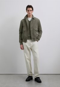 Chaqueta de pana color oliva con dos bolsillos delanteros, puesta sobre un suéter verde, combinada con pantalones blancos y zapatos negros. Fondo neutro.